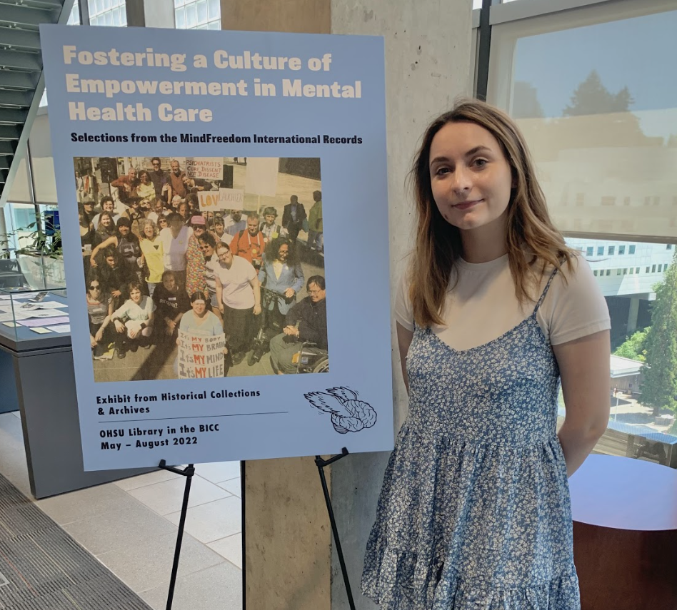 Young woman with brown hair wearing a blue dress standing in front of a blue-colored sign that reads in bold font “Fostering a Culture of Empowerment in Mental Health Care.” The sign, also features a photograph of a former protest against psychiatry.
