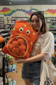 Brunette woman smiling in a craft store, holding a large "cheesy puffs" plush bag. She is wearing a white Looney Tunes shirt and denim shorts. In the background are notebooks, keychains, pins, and other craft supplies.