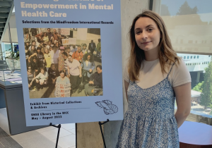 Young woman with brown hair wearing a blue dress standing in front of a blue-colored sign that reads in bold font “Fostering a Culture of Empowerment in Mental Health Care.” The sign, also features a photograph of a former protest against psychiatry.
